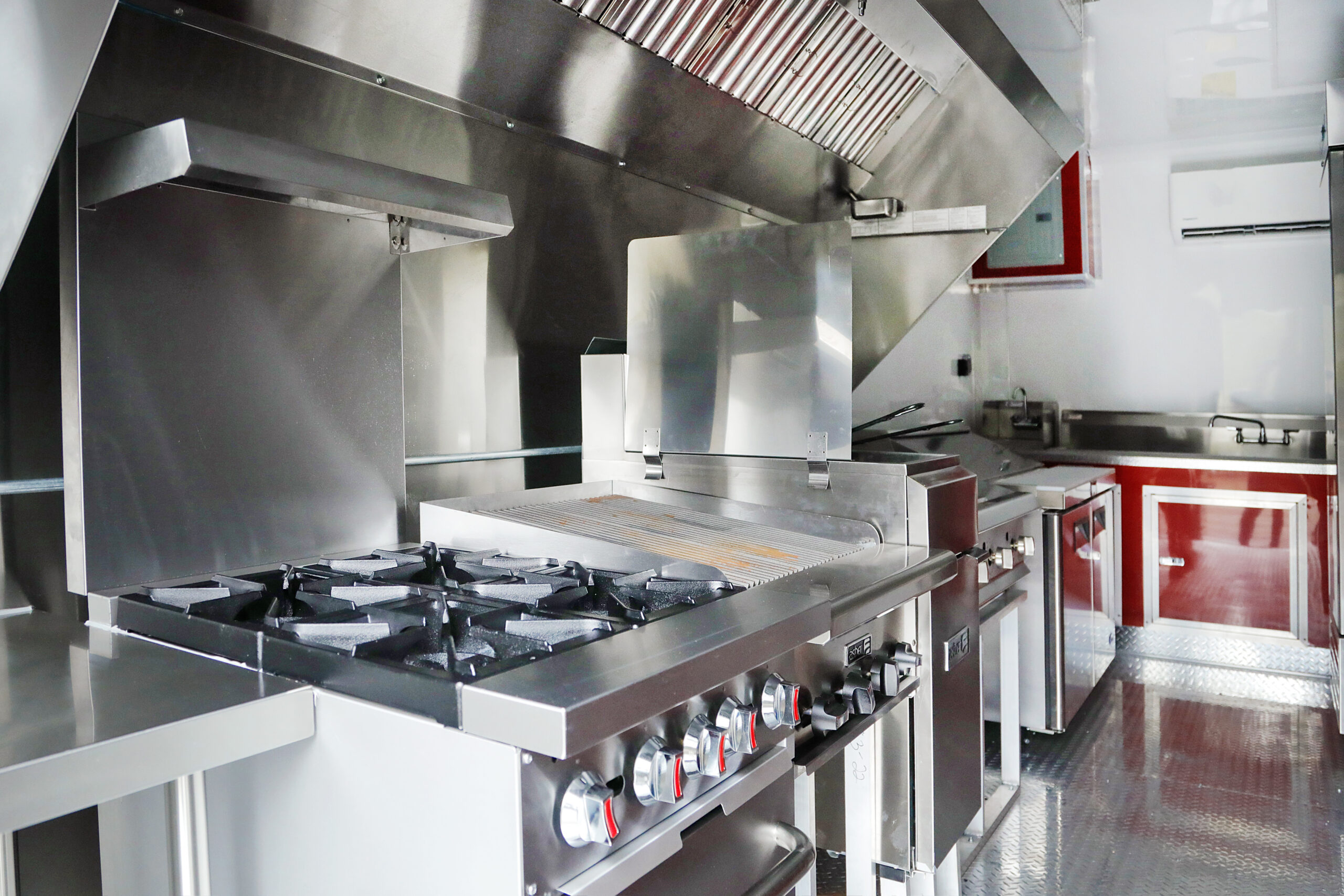A stainless steel kitchen inside a custom food trailer, with a stove, grill, and ventilation.
