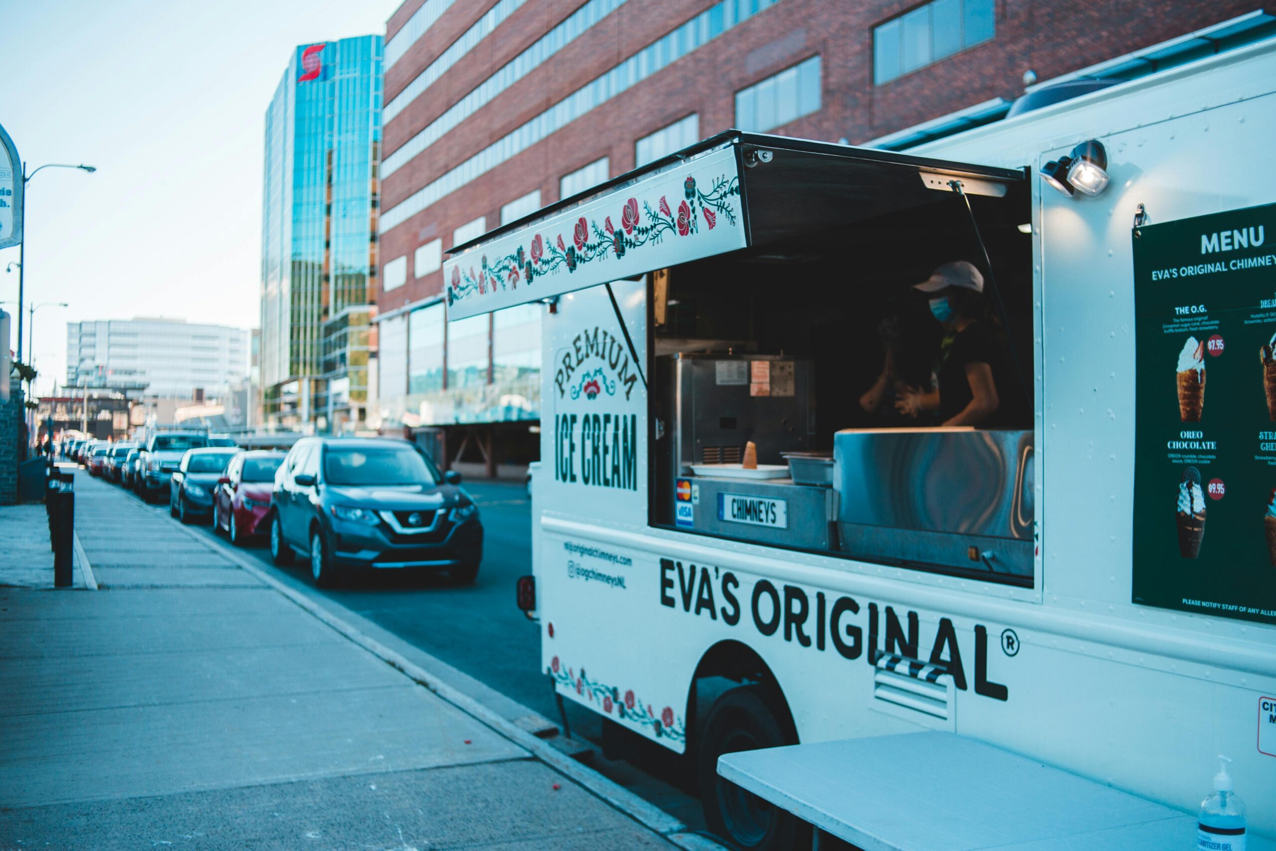 A food truck parked beside a road in front of several cars.